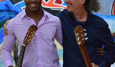 A vibrant photograph shows guitar virtuosos Ahmed Dickinson Cardenas and grammy-nomicated Eduardo Martín with their Spanish guitars smiling at each ot