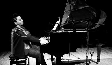 Pianist Ruth McGinley sits alone at grand piano mid performance. The dramatic image is black and white.