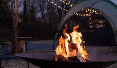 A fire burning in a firepit at dusk, with festoon lights strung through the trees at Finn Lough