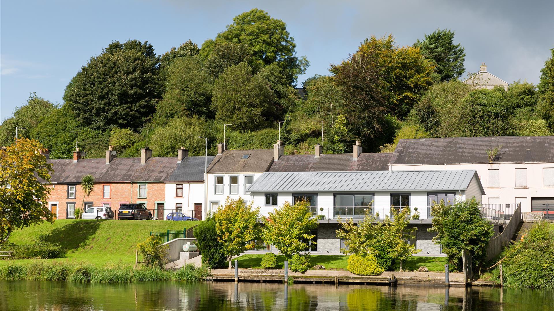 Riverside view of River Cottage with large windows and balcony, set along the water’s edge with mature trees and traditional terraced houses behind.