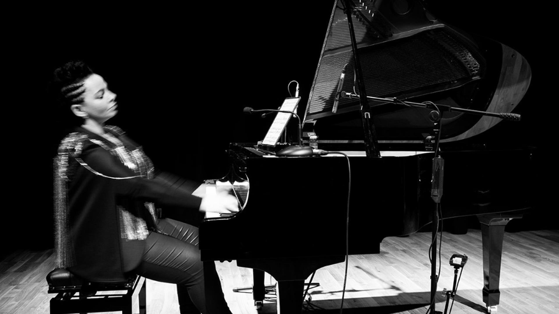 Pianist Ruth McGinley sits alone at grand piano mid performance. The dramatic image is black and white.