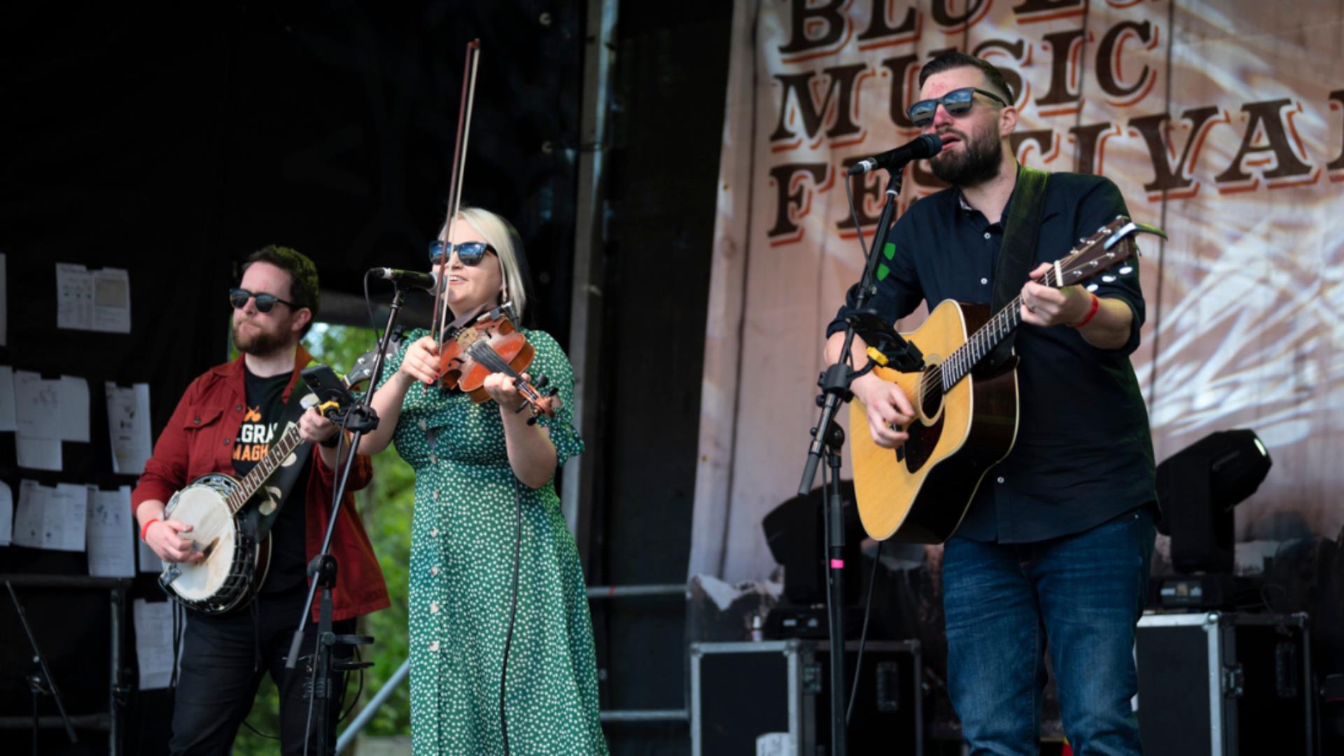 A lady in a green dress playing a violin, a man in a black tshirt playing a guitar and a man in a red shirt playing the banjo