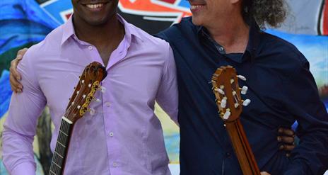 A vibrant photograph shows guitar virtuosos Ahmed Dickinson Cardenas and grammy-nomicated Eduardo Martín with their Spanish guitars smiling at each ot