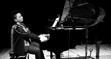 Pianist Ruth McGinley sits alone at grand piano mid performance. The dramatic image is black and white.