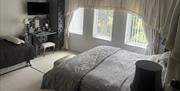 Bedroom with a large bed, a smaller bed, a vanity desk with a TV above it, and three tall windows with sheer curtains letting in natural light