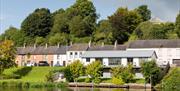 Riverside view of River Cottage with large windows and balcony, set along the water’s edge with mature trees and traditional terraced houses behind.