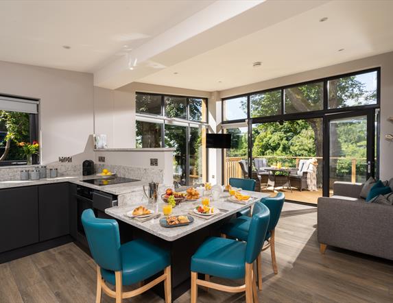 Open plan kitchen and dining area with view of lounge and outside area in background. Open patio doors floor to ceiling leading onto decked patio area